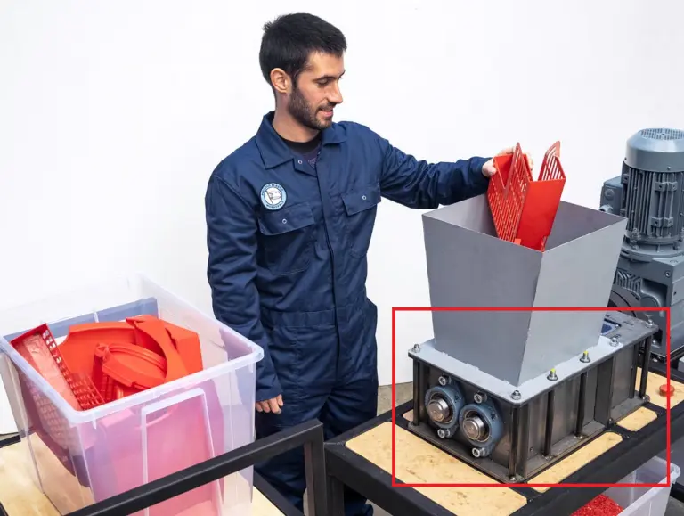Technician operating a Precious Plastic shredder machine to recycle red plastic parts in a workshop.